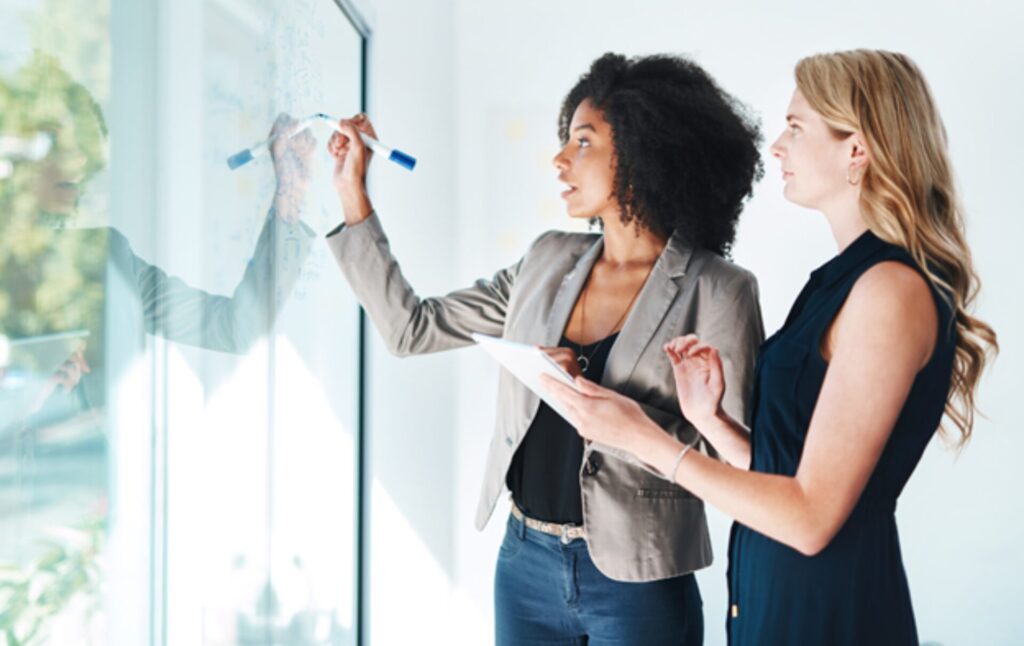 A woman speaking and drawing on a board while another watches and takes notes