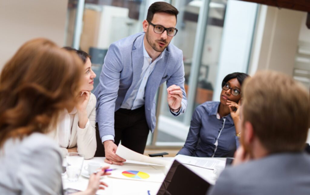 A man stands over a table in an office and speaks to a portfolio management team