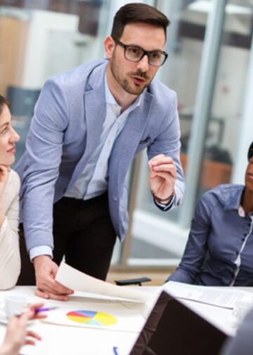 A man stands over a table in an office and speaks to a portfolio management team